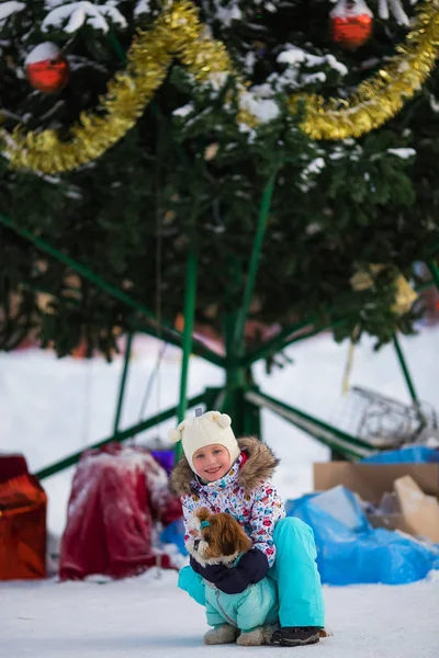 Happy little girl with a dog in overalls on a background of a Christmas tree on a winter frosty day
