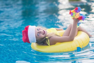 Noel Baba şapkası takan çocuk parlak güneşli bir günde mavi havuzda yüzüyor ve gülümsüyor. Mutlu yeni yıl ve noel kavramı.