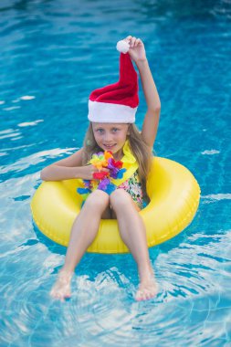 Noel Baba şapkası takan çocuk parlak güneşli bir günde mavi havuzda yüzüyor ve gülümsüyor. Mutlu yeni yıl ve noel kavramı.