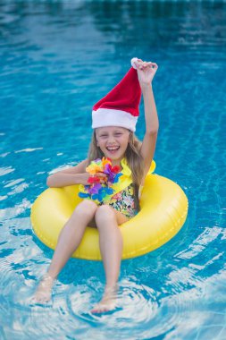 Noel Baba şapkası takan çocuk parlak güneşli bir günde mavi havuzda yüzüyor ve gülümsüyor. Mutlu yeni yıl ve noel kavramı.