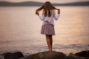 Arkadan bak. Göldeki bir kayanın üzerinde duran uzun saçlı tatlı kız. Tatillerde uyum, huzur ve mutluluk kavramları..