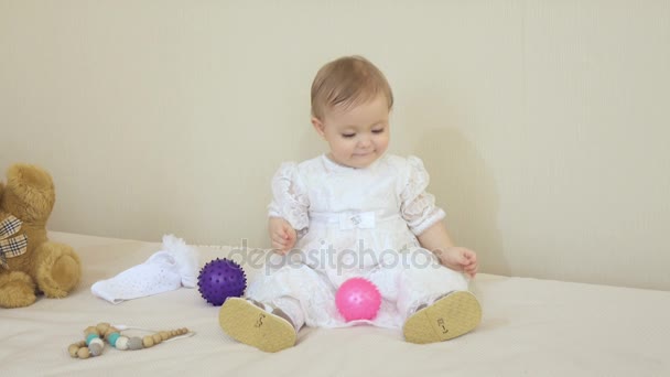 En liten leende flicka i en vit klänning spelar färgglada bollar — Stockvideo