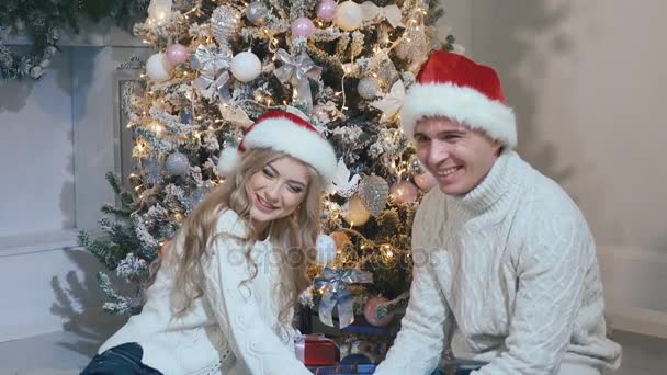 Ένας νεαρός άντρας και ένα όμορφο κορίτσι κάθονται κοντά το ντυμένοι χριστουγεννιάτικο δέντρο — Αρχείο Βίντεο