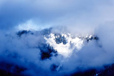 Sun shining through the clouds on the Snow Capped Peak of Coquitlam Mountain in the Coast Mountain Range seen from the shore of Pitt Lake in the Fraser Valley of British Columbia, Canada clipart