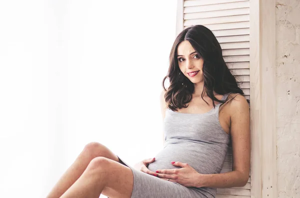 Mulher grávida em vestido cinza sentado perto da janela — Fotografia de Stock