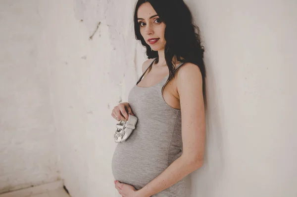 Kobieta w ciąży w pobliżu ściany i gospodarstwa buty dziecięce — Zdjęcie stockowe