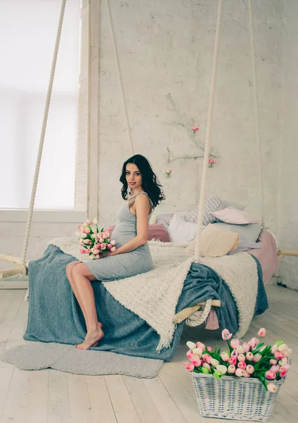 Yatağın üstüne oturan gri elbiseli mutlu hamile kadın — Stok fotoğraf