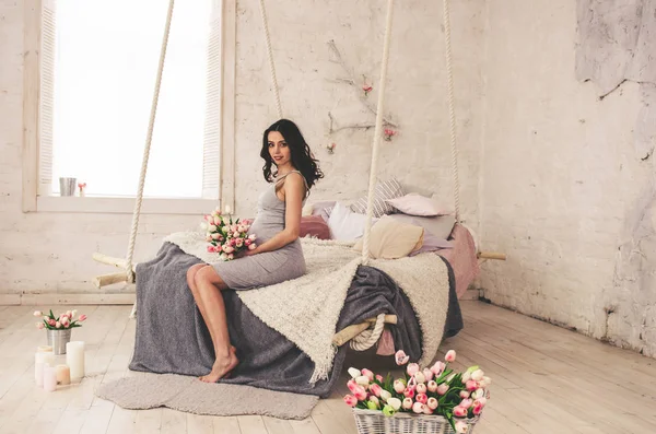 Glad gravid kvinna i grå klänning sitter på sängen — Stockfoto