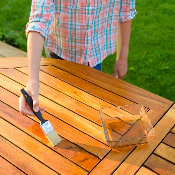 Mano sosteniendo un pincel aplicando pintura de barniz sobre una mesa de jardín de madera —  Fotos de Stock