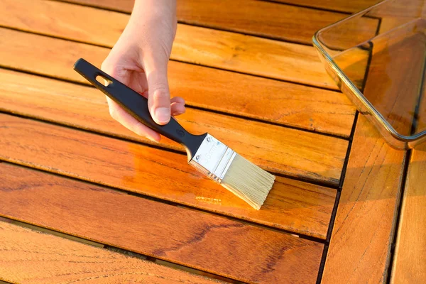 Hand holding a brush applying varnish paint on a wooden garden table — Stock Photo, Image