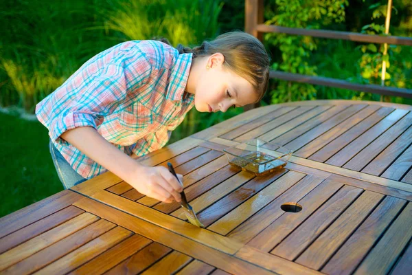 Menina segurando uma escova aplicando tinta de verniz em uma mesa de jardim de madeira — Fotografia de Stock