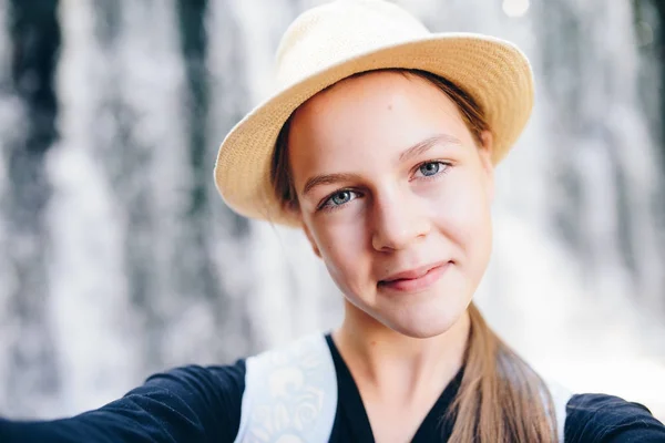 Молода дівчина в капелюсі бере селфі на свій смартфон з видом на водоспад — стокове фото