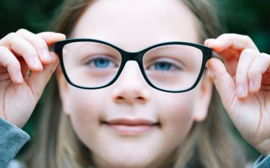 Closeup portrait of little girl  with myopia correction glasses. Girl is holding her eyeglasses right in front of camera with two hands - focus on glasses - shallow depth of field clipart
