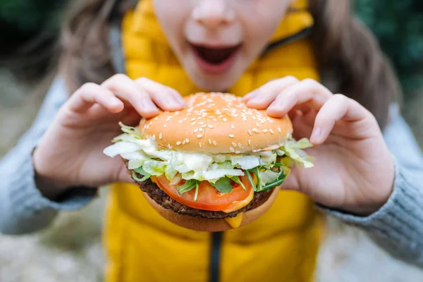 Küçük kız domates, marul, roka, sığır eti ve sosla büyük bir çizburger yiyor. İki elle tutuyor. Saha derinliği sığ.
