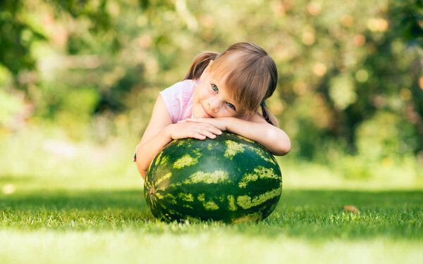Маленькая девочка, сидящая на корточках у большого арбуза на газоне в летнем саду - неглубокая глубина поля
