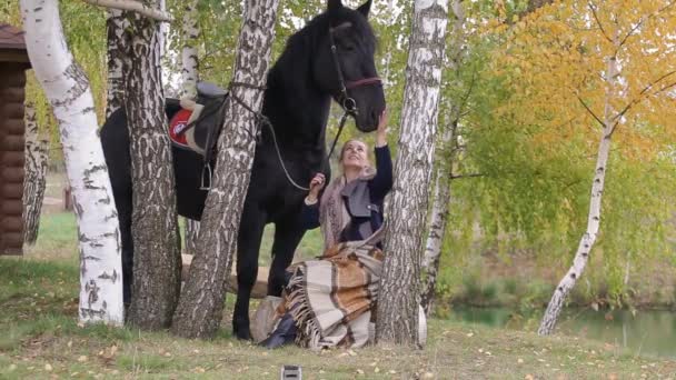 Menina sentada em um banco entre as bétulas no outono com um cavalo — Vídeo de Stock