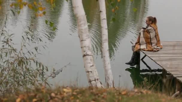 Mulher menina sentar-se no banco de madeira sob o ramo de salgueiro mover-se no vento perto do lago — Vídeo de Stock
