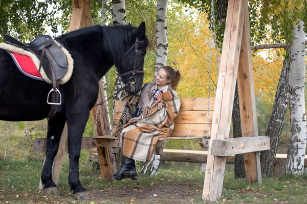 Κορίτσι σε ένα καρό με ένα μαύρο άλογο το φθινόπωρο υπό ένα σημύδα δέντρο σε ένα παγκάκι — Φωτογραφία Αρχείου