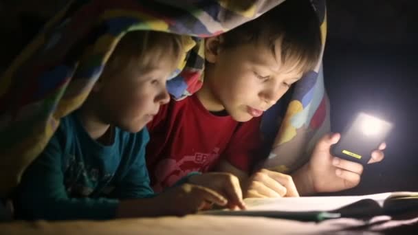 A gyermekek olvasni egy könyvet, egy egy zseblámpával takaró alatt — Stock videók