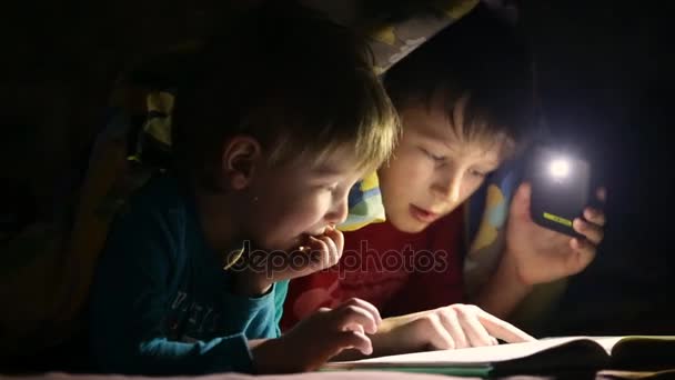 Дети читают книгу под одеялом с фонариком — стоковое видео