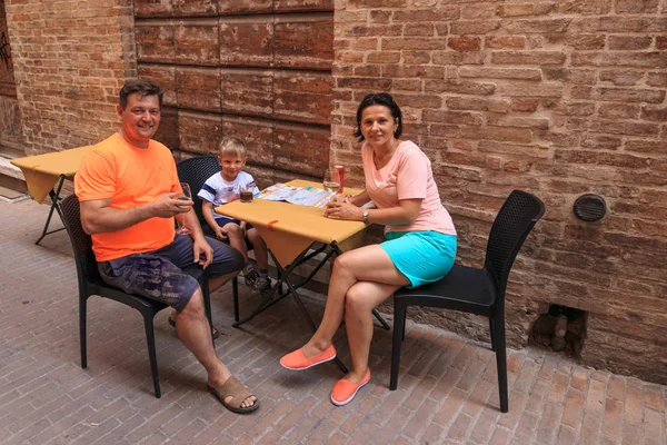 La familia se sienta en una mesa de café en la estrecha calle de Italia . — Foto de Stock