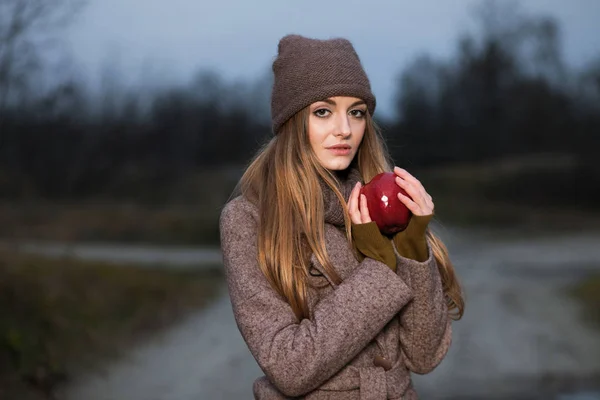 Meisje in een gebreide cap en een sjaal met een appel in de herfst op aard. — Stockfoto