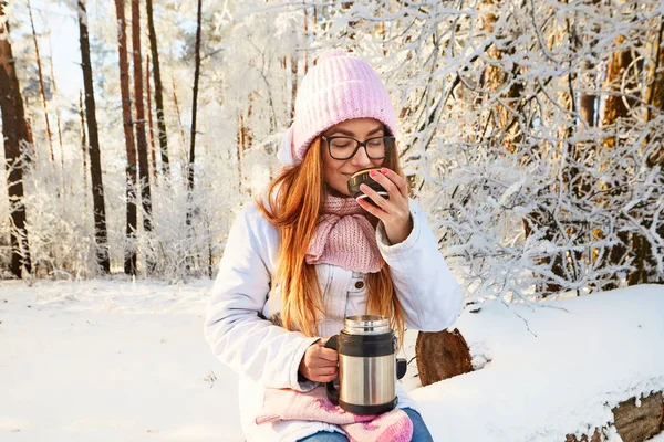 Menina em um chapéu rosa com uma garrafa térmica no inverno na floresta . — Fotografia de Stock