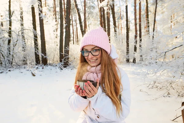 Menina em um chapéu rosa bebendo chá de uma garrafa térmica no inverno na floresta . — Fotografia de Stock