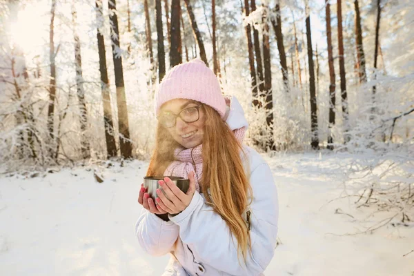 Menina em um chapéu rosa bebendo chá de uma garrafa térmica no inverno na floresta . — Fotografia de Stock