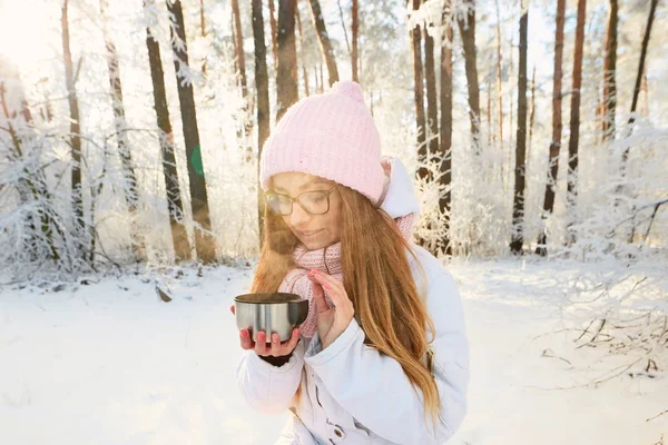 Menina em um chapéu rosa bebendo chá de uma garrafa térmica no inverno na floresta . — Fotografia de Stock