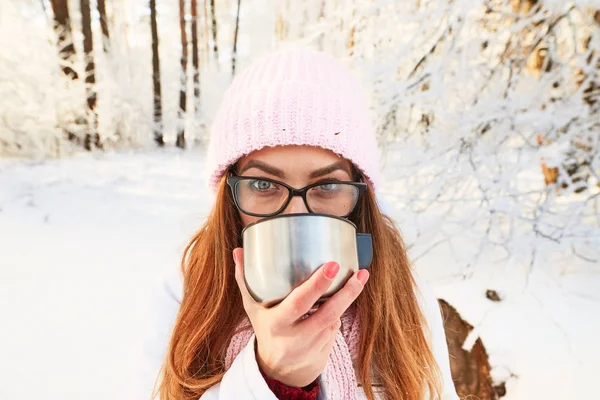Lány rózsaszín kalapot teát iszik egy Termosz télen az erdő. — Stock Fotó