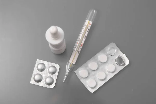 Medizinisches Thermometer mit Tabletten und Nasenspray auf grauem Hintergrund im Studio. — Stockfoto