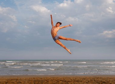 Sahilde atletik bir genç, denizin ve gökyüzünün arka planında zıplıyor.