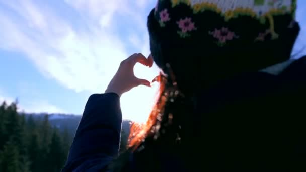 Krásná dívka ukazuje gesta srdce na pozadí oblohy a slunce poblíž zimní les — Stock video