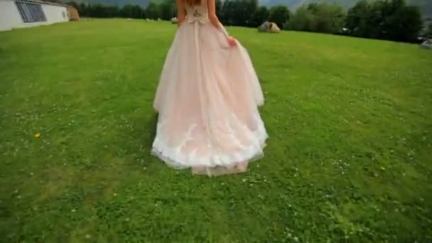 Die rothaarige Braut in einem verschwenderischen Kleid schreitet im grünen Gras vor der Kulisse der hohen Berge. Hochzeitstag — Stockvideo