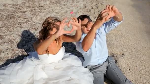 Счастливые влюбленные молодожены делают сердце из пальцев на пляже — стоковое видео