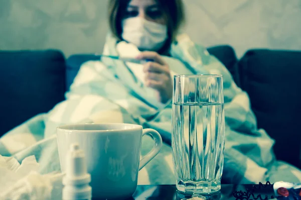 風邪や季節性インフルエンザの概念 マスクの少女は 薬でテーブルの近くの毛布に包まれたソファに座っている温度計を見ています 熱い癒しの飲み物を飲む — ストック写真