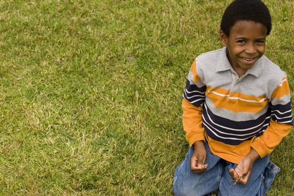 Bonito menino afro-americano sentado na grama fora . — Fotografia de Stock