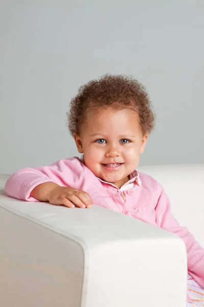 Linda niña de raza mixta sonriendo sentada en un sofá . — Foto de Stock