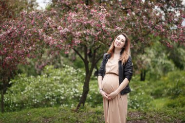 Baharda çiçek açan ağacın yanında duran bej rengi elbise ve siyah sabun paltosu giymiş, gülümseyen hamile bir kadın. yatay portre