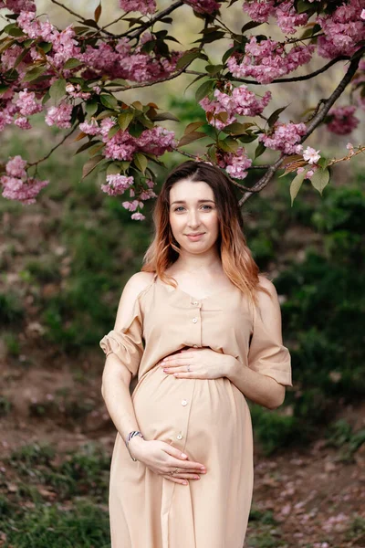 春天，身穿米黄色衣服的年轻孕妇站在开花的树上。垂直肖像 — 图库照片