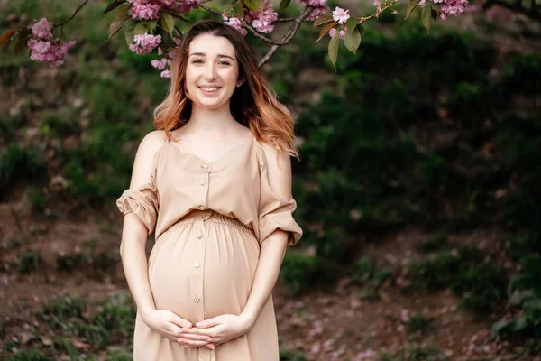 Fiatal, mosolygó terhes nő bézs ruhában áll a virágzó fa mellett tavasszal. horizontális portré — Stock Fotó
