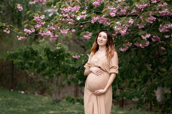 Fiatal, mosolygó terhes nő bézs ruhában és fekete laposkabátban áll a virágzó fa mellett tavasszal. horizontális portré — Stock Fotó