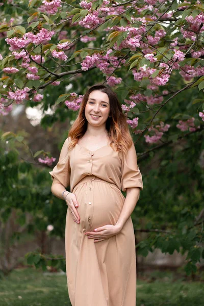 Fiatal terhes nő bézs ruhát visel, tavasszal virágzó fa mellett áll. függőleges portré — Stock Fotó