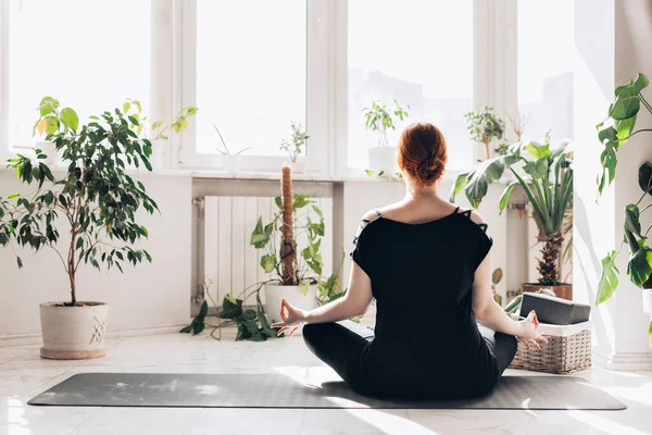 Mujer pelirroja caucásica practica yoga en casa en una sala de estar de luz blanca. ventana y plantas fondo —  Fotos de Stock