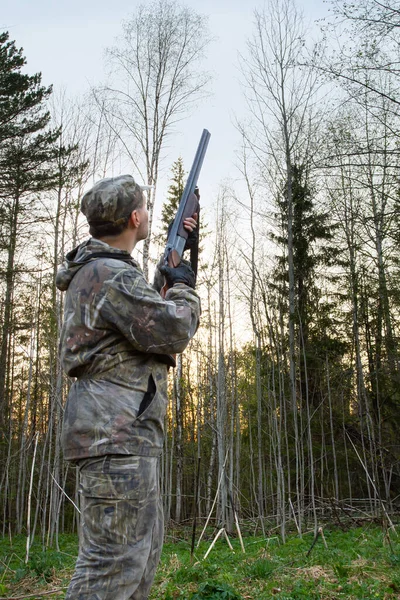A hunter with a rifle in his hands stands in the evening in a forest clearing and waits for the appearance of a woodcock, which makes a courtship flight (roding)
