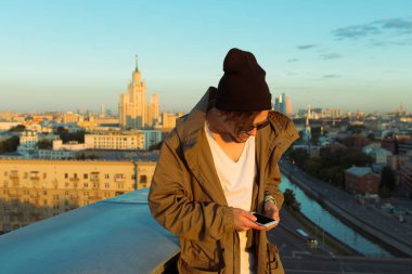 MOSCOW, RUSSIA - 13 Haziran 2016: Yüksek çatının kenarında oturan genç ve cesur adam yaz aylarında Moskova 'nın şafak vakti iPhone ve çarpıcı manzarasına benzeyen telefona bakıyor