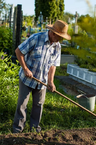 An elderly rural man in a straw hat is working near the grave in the cemetery.