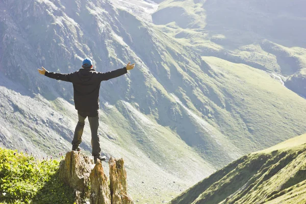 Homem de jaqueta preta em pé sobre pedra com as mãos para cima acima montagens ao pôr do sol — Fotografia de Stock