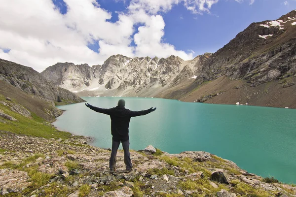 Man in black jacket standing with hands-up above blue lake in mountains — Stock Photo, Image